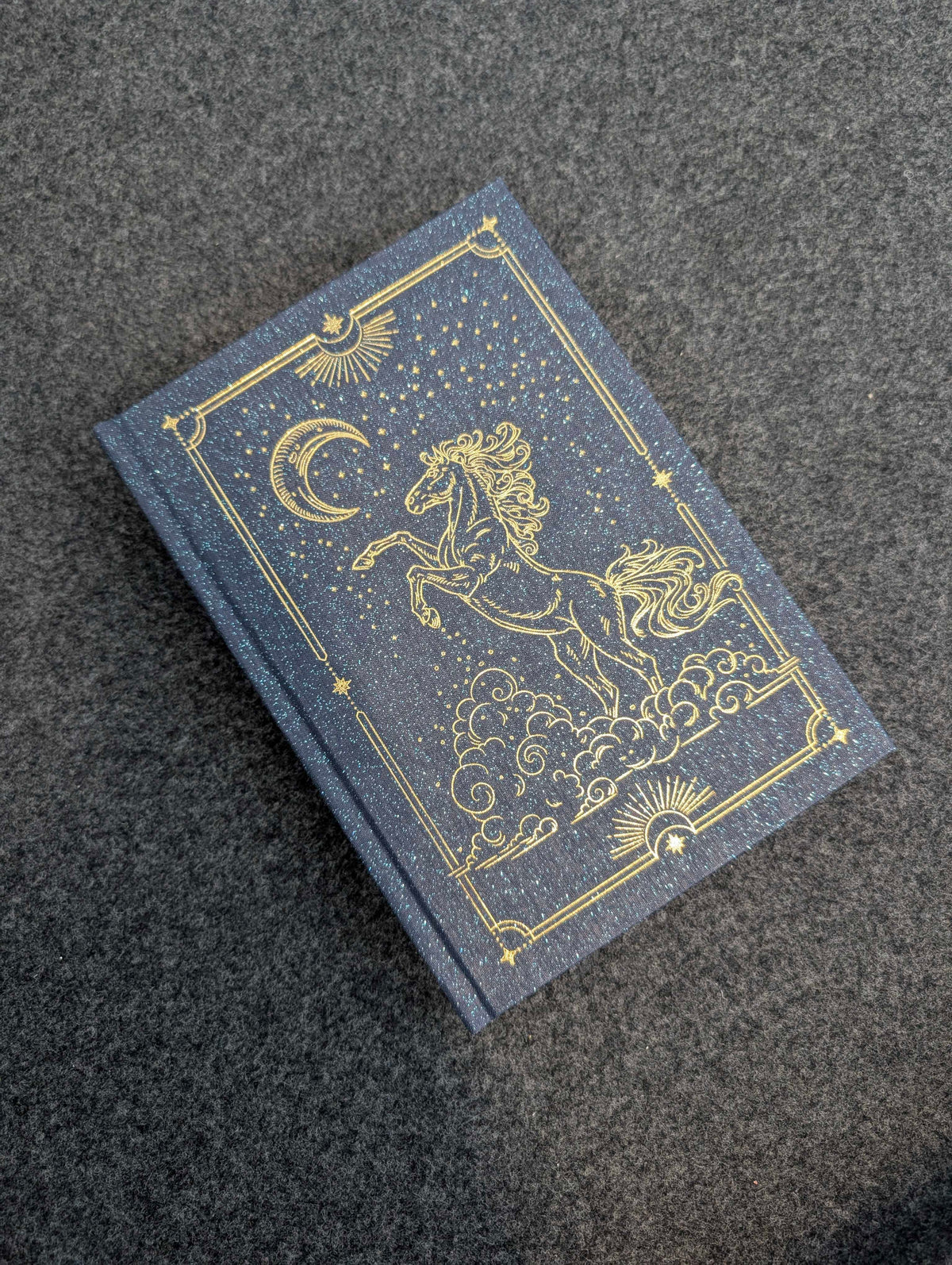 Blue book with gold illustrations of a horse and celestial elements on a dark textured background #color_Midnight Stars