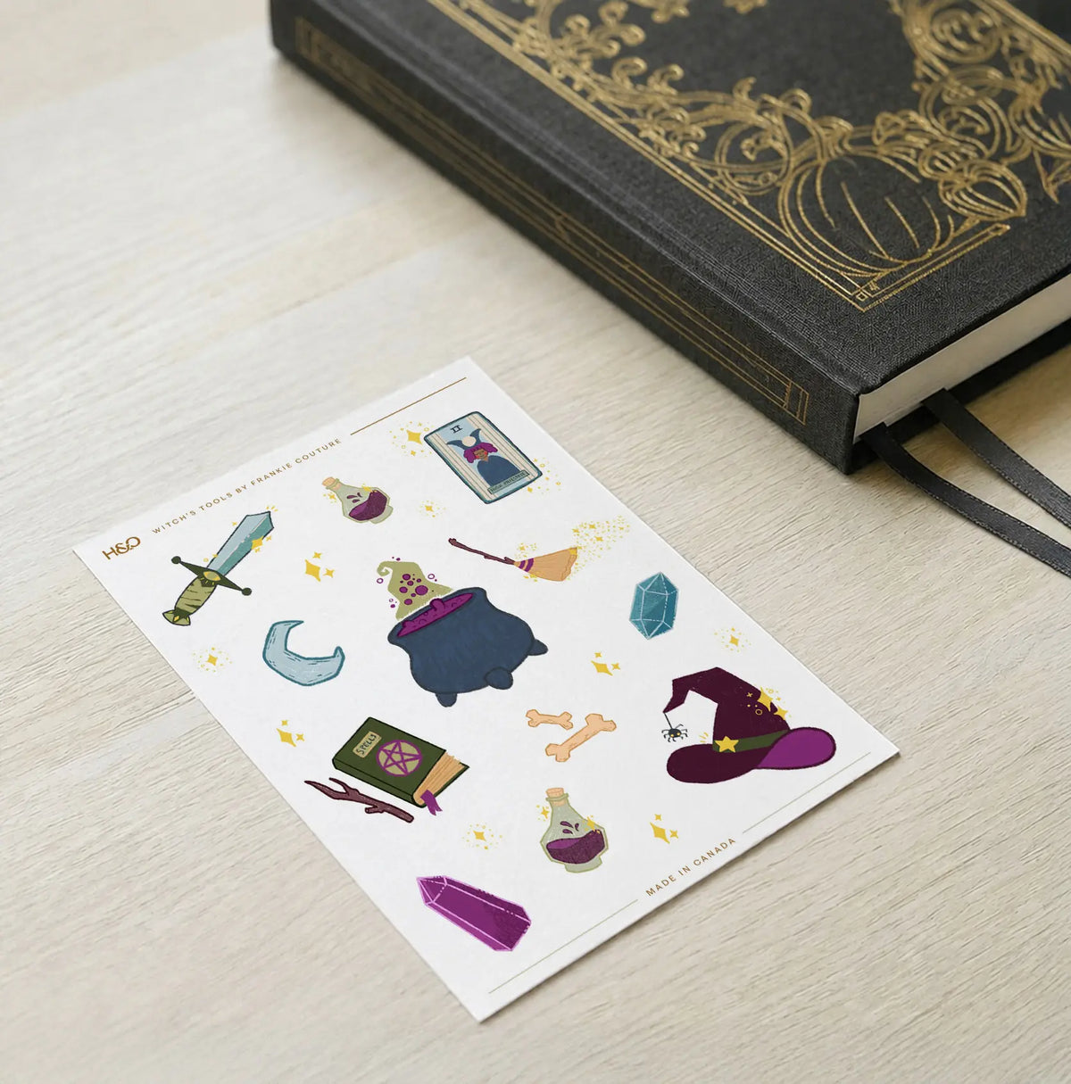 Witch's Tools sticker sheet by Frankie Couture on a pale wood desk beside a navy linen notebook with a gold rabbit emblem and blue ribbon markers